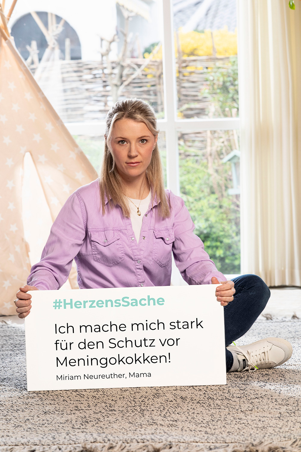 Miriam Neureuther sitzt in einem Kinderzimmer mit einem Schild vor sich auf dem Boden, eine Kampagne für Meningokokkenimpfung
