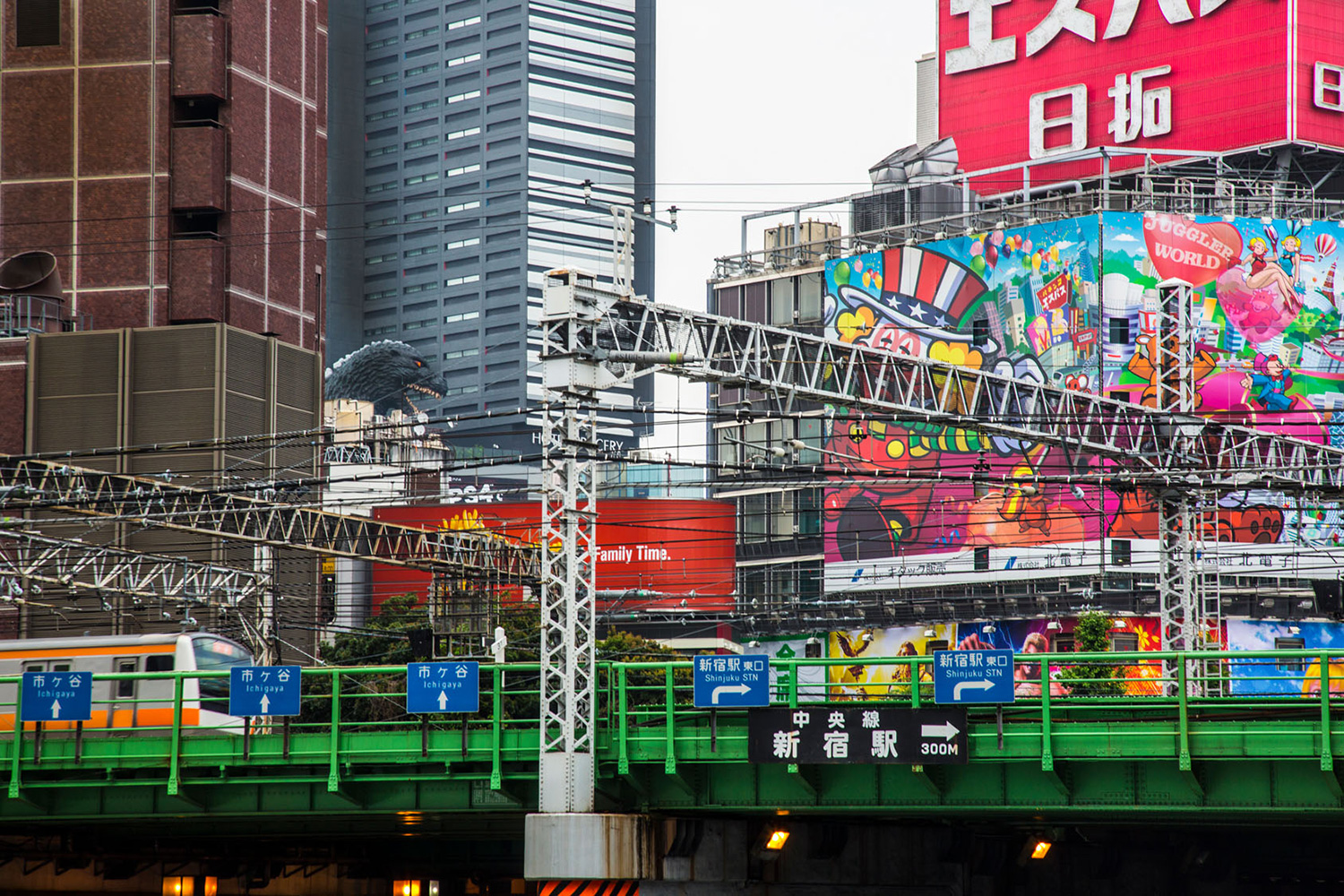 Eine Aufnahme von Häusern und einer S-Bahn in Tokio, im Hintergrund sieht man Godzilla, überall bunte Werrbung und Schriftzüge