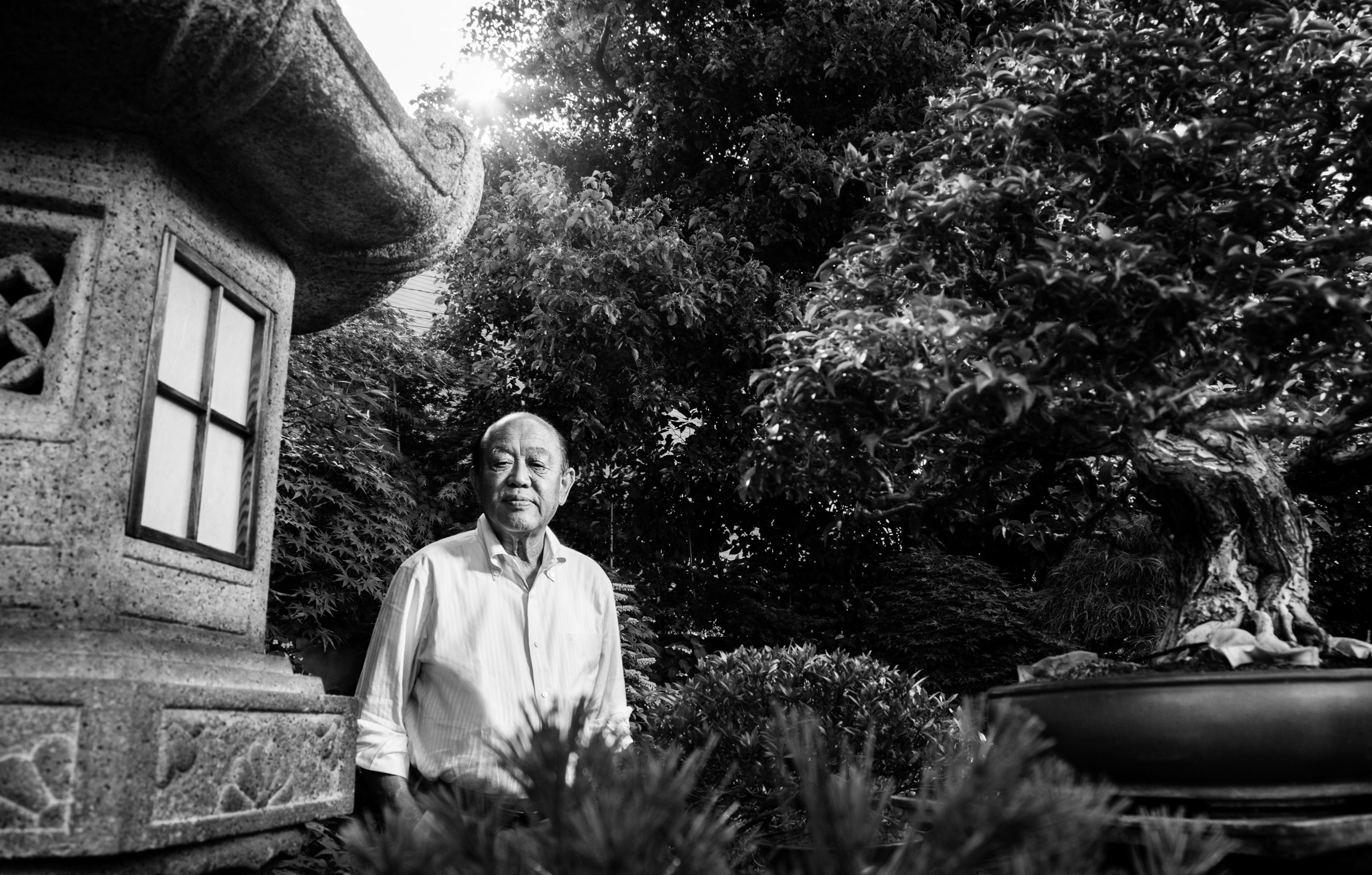 Portrait eines berühmten japanischen Bonsi Meisters in seinem Garten hinter einem Steinhaus zwischen Bonsai Bäumen
