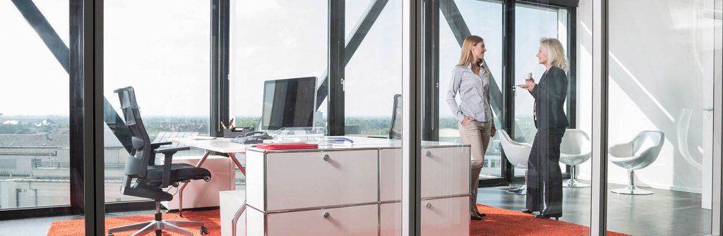 Zwei Frauen stehen in einem großen hellen Büroraum mit großen Fenstern nach außen