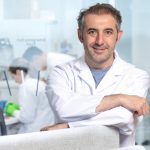 Portrait von Ali Ertürk vor einer Glaswand hinter der sich ein Labor mit Mitarbeitern zeigt