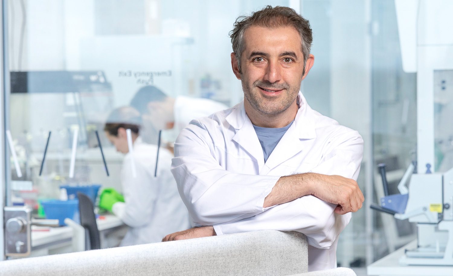 Portrait von Ali Ertürk vor einer Glaswand hinter der sich ein Labor mit Mitarbeitern zeigt
