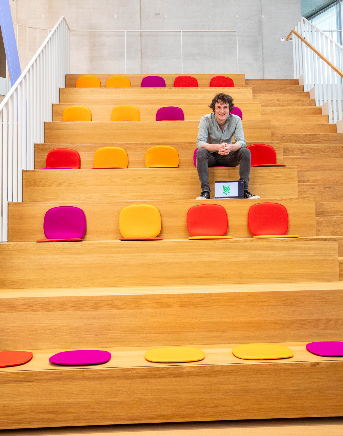 Ein Mann sitzt auf den Stufen einer Aula mit Holzboden und bunten Sitzauflagen, ein Laptop steht vor ihm und er schaut in die Kamera