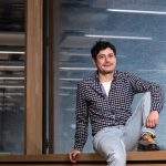 Ein junger Wissenschaftler sitzt bequem auf einem Fensterbrett eines großen Fensters mit einer großen markise und den Refelxen von Lampen im Hintergrund