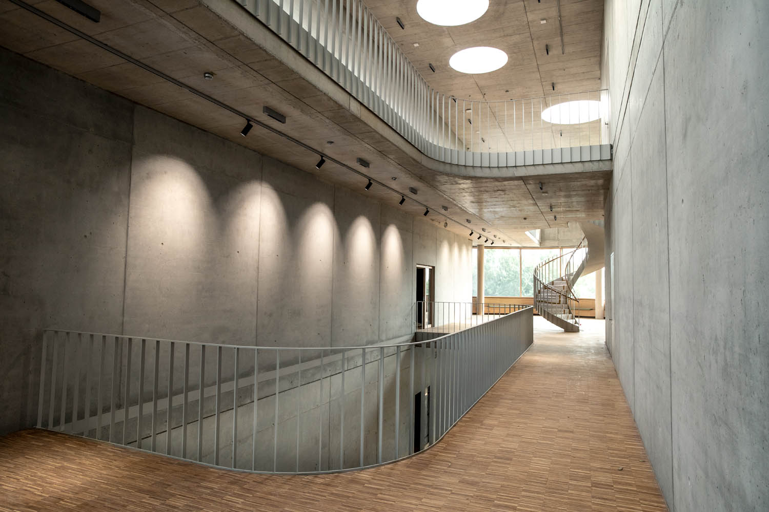 Gang in einem großen Gebäude, die Wände sind roh Beton, der Boden Parkett am Ende des Ganges sieht man eine Wendeltreppe