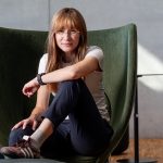 Eine Frau mit Brille und langen roten Haaren sitzt in einem grünen Sessel ihr Arm ist aufgestützt sie schaut direkt in die Kamera