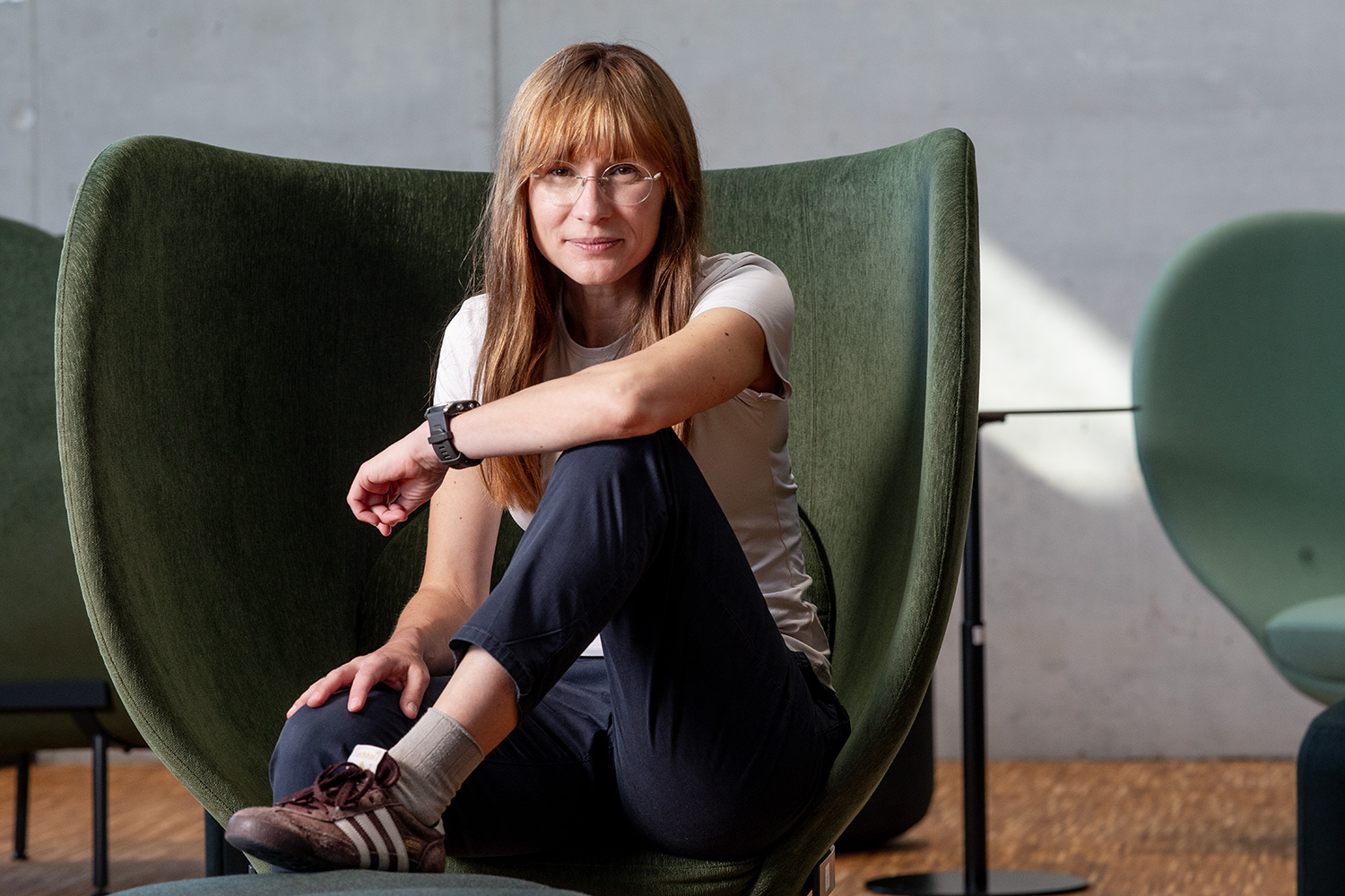Eine Frau mit Brille und langen roten Haaren sitzt in einem grünen Sessel ihr Arm ist aufgestützt sie schaut direkt in die Kamera