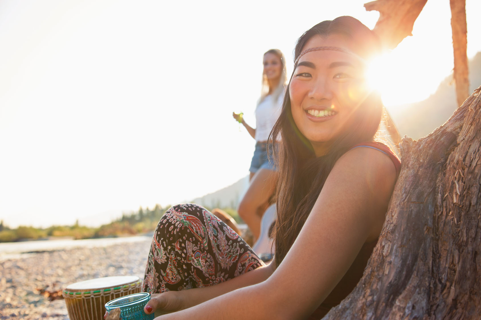 Eine junge Frau lächelt in die kamera, das Sonnenlicht scheint von hinten und sie sitzt angelehnt an einen baum in der natur