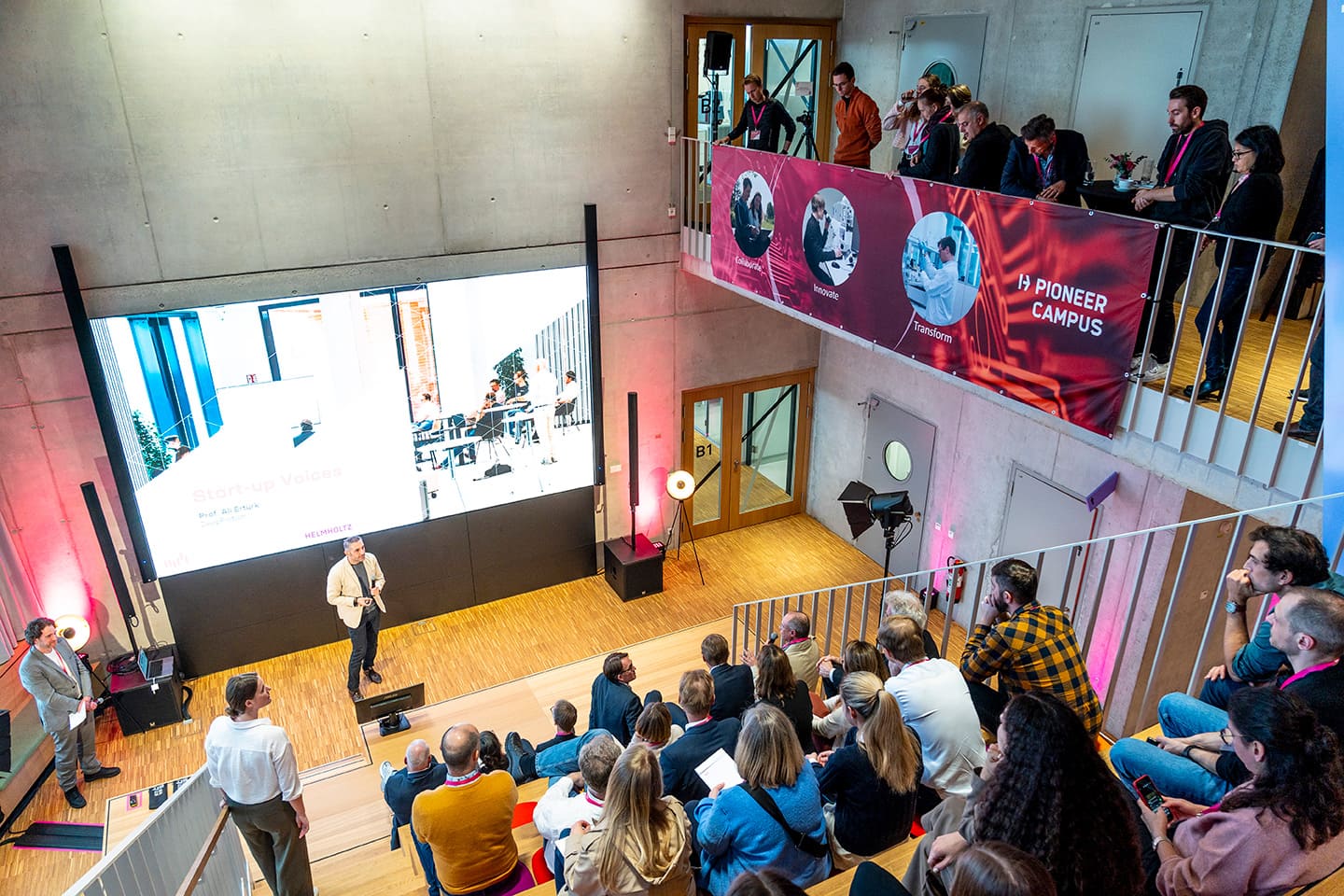Vortrag eines Mannes vor einer Videowand, spricht zu dem Publikum auf den Stufen einer großen Aula